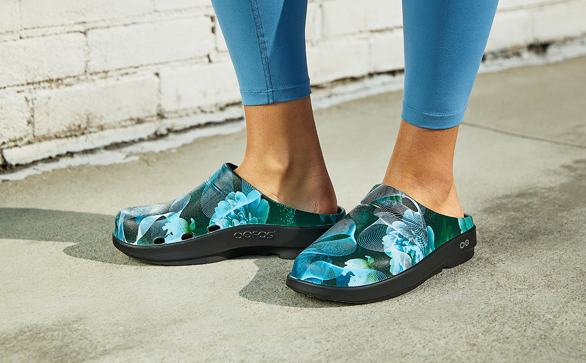 Person wearing floral clogs on a concrete surface with a white brick wall in the background | lifestyle