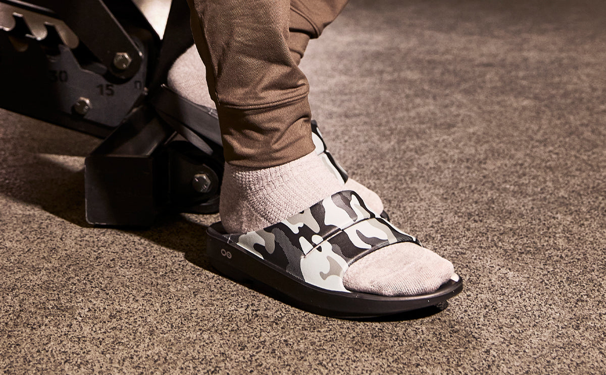 A close-up of a person's foot wearing a black and gray camouflage slide sandal with a light beige textured toe cap and a light gray sock, standing on a textured brown carpet | lifestyle