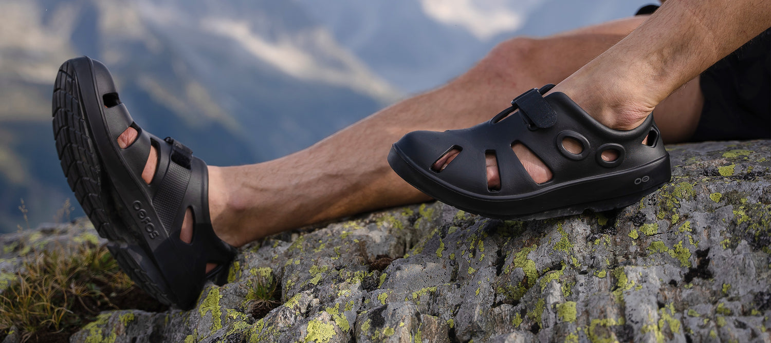 Man wearing Black OOcandoo recovery sandals sitting on cliffside