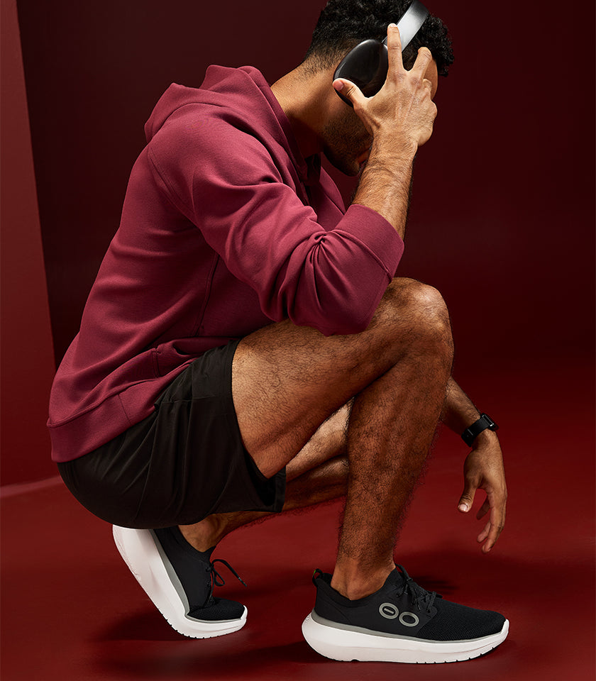 A man in a maroon hoodie and black athletic shorts crouches while wearing black sneakers with white soles and black headphones, set against a dark red studio background.