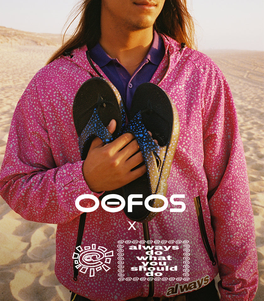 A person standing in a sandy desert holds a pair of black OOFOS flip-flops with blue and gold patterned straps. The person is wearing a pink patterned zip-up hoodie over a purple collared shirt.