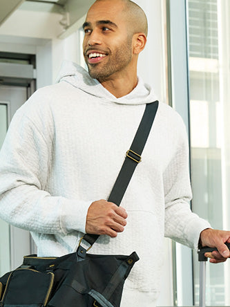 Man in a white hoodie with a black bag over his shoulder, standing indoors.