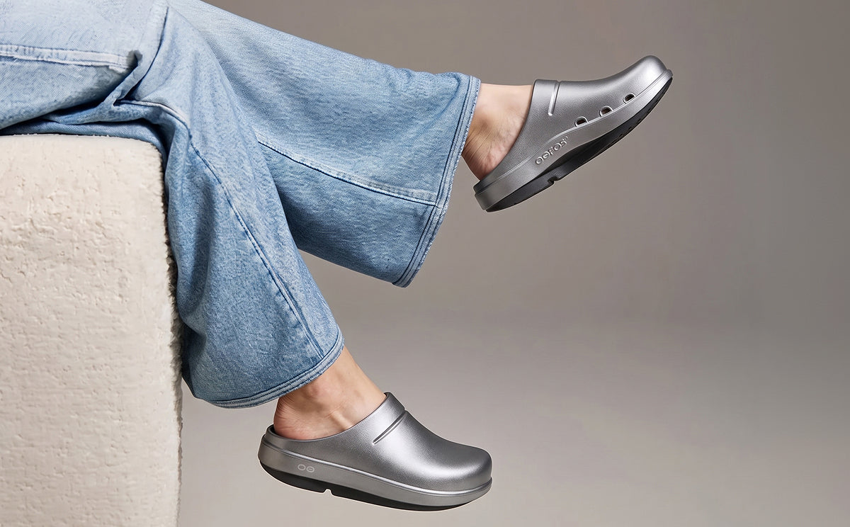 A person wearing metallic silver clogs with wide-leg light blue jeans, sitting on a cream-colored textured chair against a neutral gray background | lifestyle