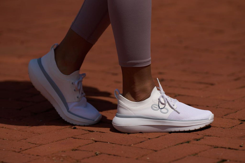 White athletic sneakers with light gray accents worn by a person standing on a red brick surface.