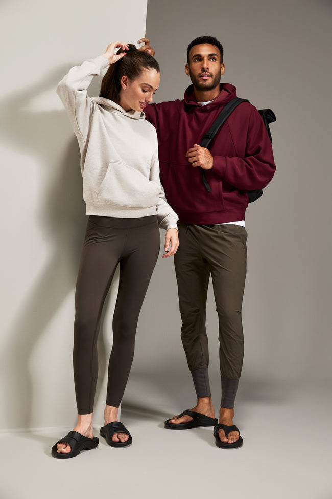 A man and a woman modeling casual apparel in a studio setting. The woman wears a light beige hoodie and dark brown leggings, while the man wears a burgundy hoodie, olive green joggers, and black sandals, with a black backpack strap visible.