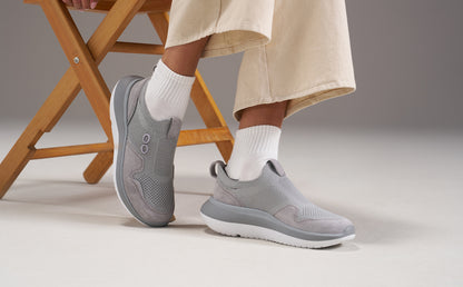 A person wearing light gray slip-on sneakers with white crew socks and beige trousers, seated on a wooden folding chair against a neutral studio background | lifestyle