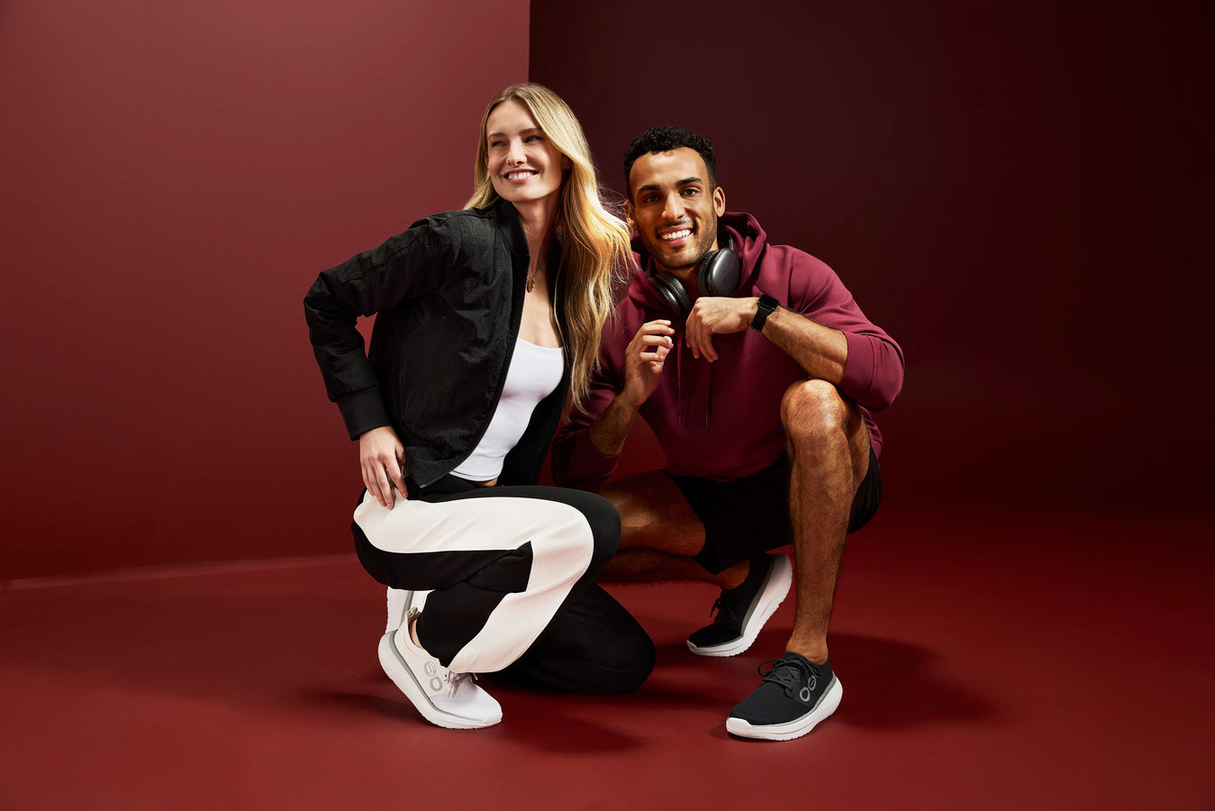A man and a woman pose together in a studio setting with a dark red background. The woman wears a black blazer over a white top with black and white track pants and white sneakers.