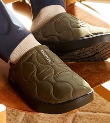 Green quilted slippers with Oofos branding worn indoors on a wooden floor.