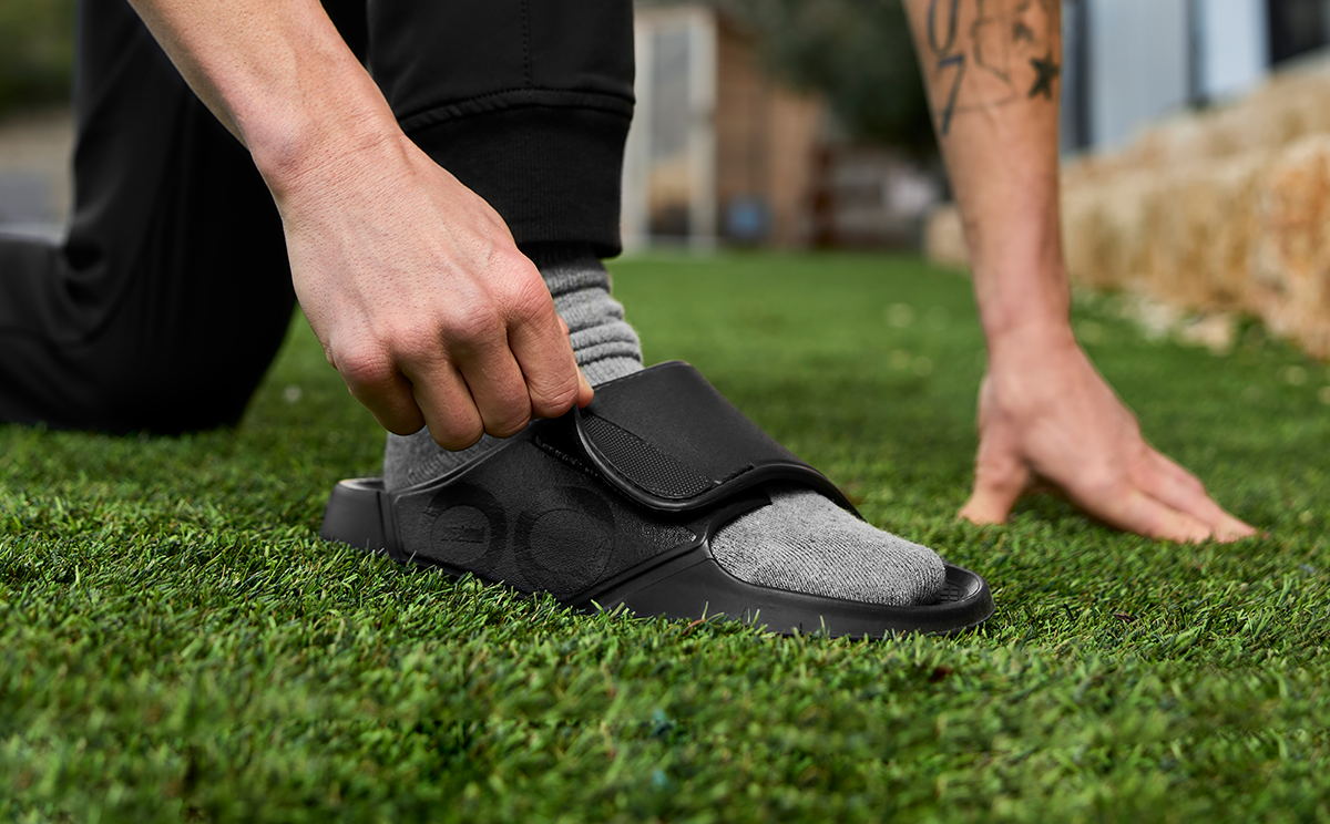 Men's OOahh Sport Flex Sandal - Matte Black close up of model adjusting strap of shoe kneeling on grass | lifestyle