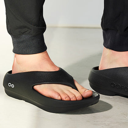 Person wearing black sandals with Oofos branding on a light background