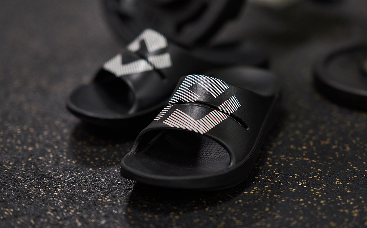 FJ X OOFOS Men's OOahh Sport Slide Sandal - Black close up of shoes on gym floor | lifestyle