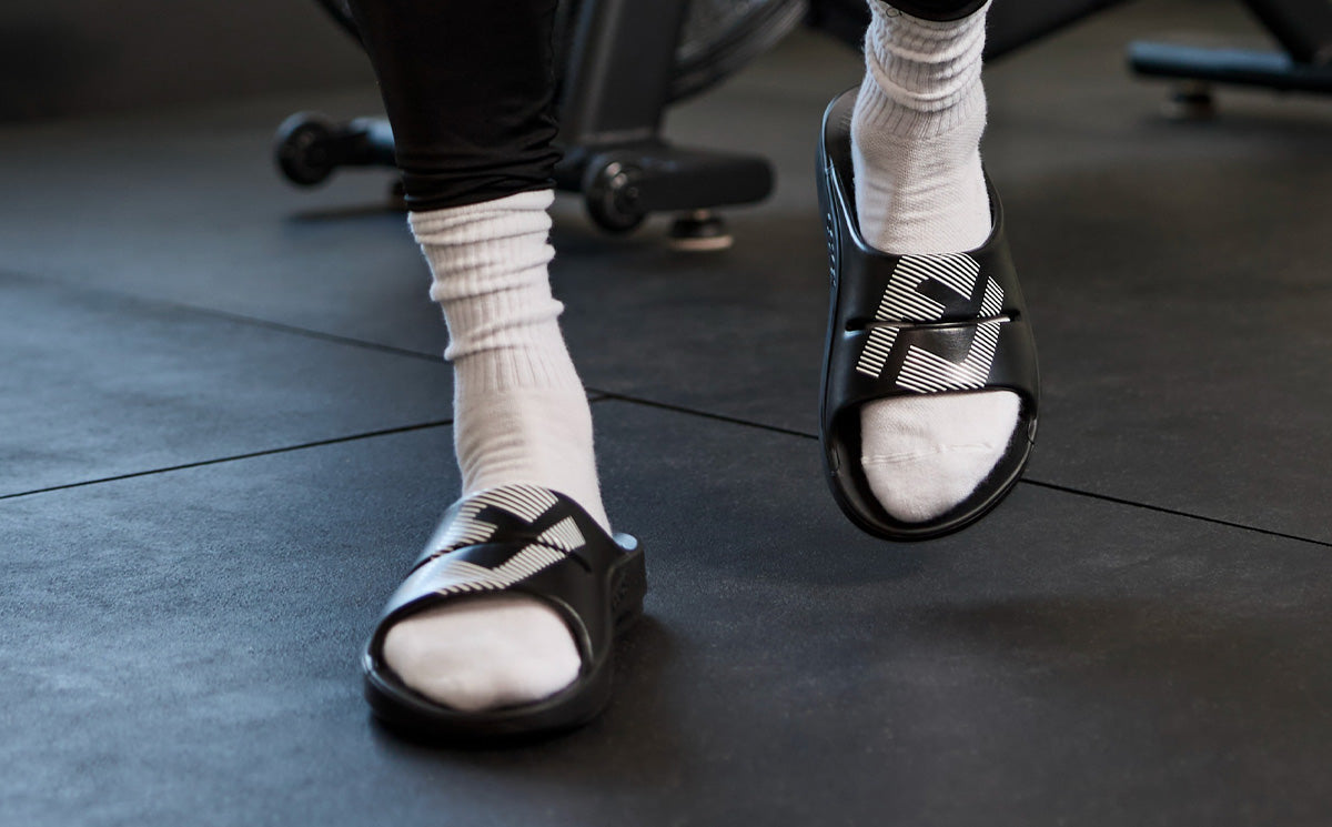 FJ X OOFOS Men's OOahh Sport Slide Sandal - Black front view of model walking in shoes in gym | lifestyle