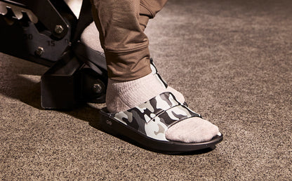 A close-up of a person's foot wearing a black and gray camouflage slide sandal with a light beige textured toe cap and a light gray sock, standing on a textured brown carpet | lifestyle