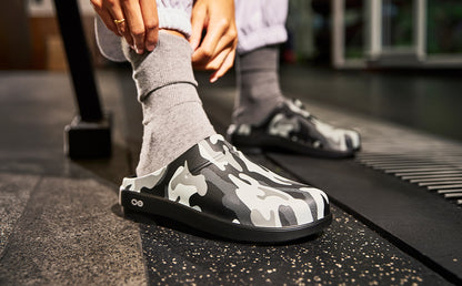 A person wearing gray socks and black-and-white camouflage slip-on clogs in a gym setting, with one shoe being adjusted and the other visible in the background | lifestyle
