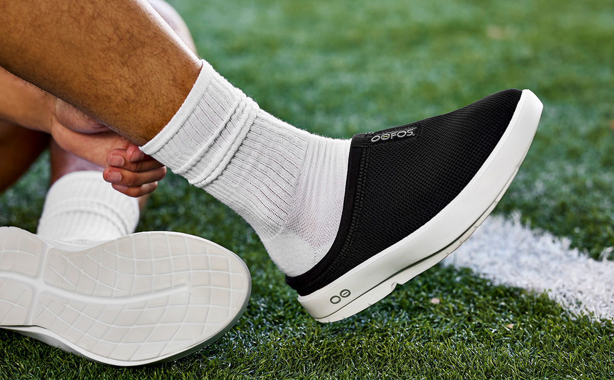 Men's OOcoozie Sport Mule - White Black close up of shoe on model sitting on field | lifestyle