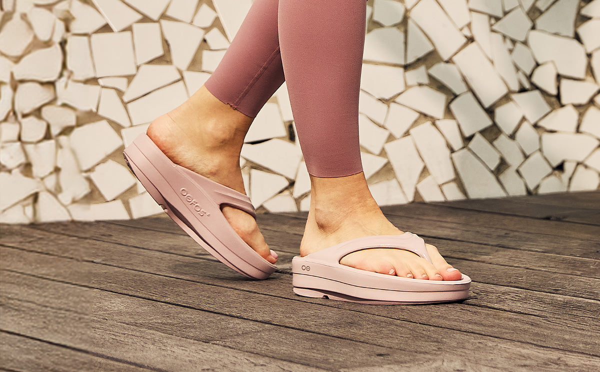 Person wearing pink sandals on a wooden deck with a mosaic stone wall in the background | lifestyle