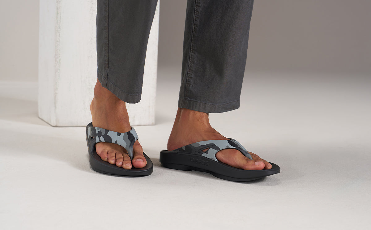 A person wearing gray pants and black flip-flop sandals with a gray camouflage pattern on the straps, standing on a light gray studio floor | lifestyle