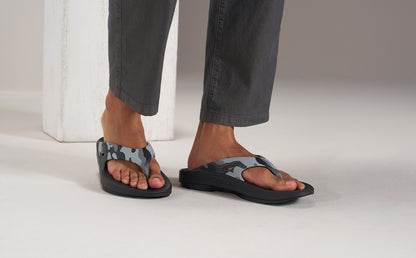 A person wearing gray pants and black flip-flop sandals with a gray camouflage pattern on the straps, standing on a light gray studio floor | lifestyle