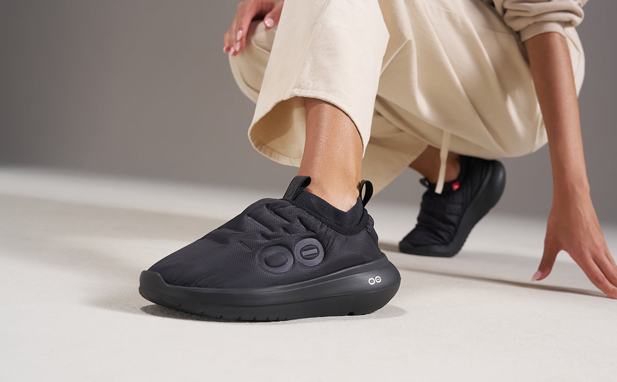A person wearing black slip-on sneakers and beige pants, crouching in a studio setting with a neutral gray background | lifestyle