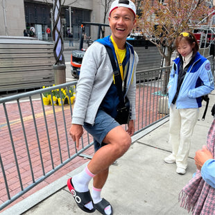 Person in a gray jacket and blue shorts posing on a city sidewalk with another person in the background.