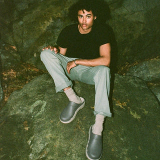 A man sits on a mossy rock in a dark outdoor setting, wearing a black t-shirt, light gray pants, white socks, and gray slip-on shoes.