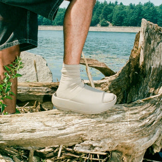 A person wearing a light beige slip-on platform sandal with a ribbed ankle sock, standing on a large weathered log outdoors near water and trees.