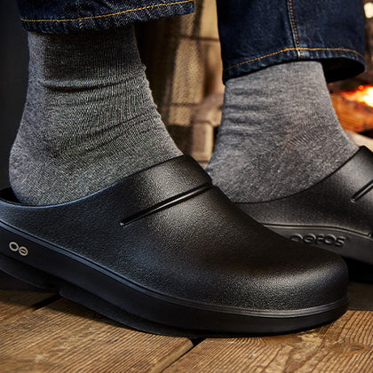 Black slip-on shoes worn with gray socks on a wooden floor.