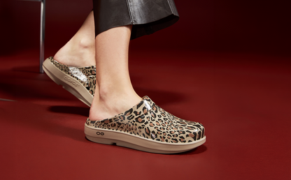 A person wearing leopard-print clogs with beige soles, shown from the calves down against a dark red studio background | lifestyle
