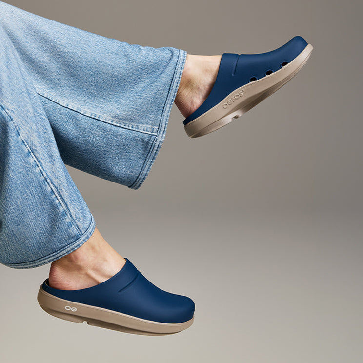 A person wearing blue clogs with light beige soles, sitting on a cream-colored chair. The person is also wearing light blue jeans, set against a neutral gray studio background.