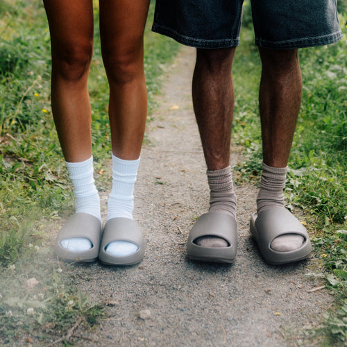 Two people standing on a dirt path outdoors, shown from the waist down. One wears white socks and taupe slide sandals, and the other wears gray socks and matching taupe slide sandals.