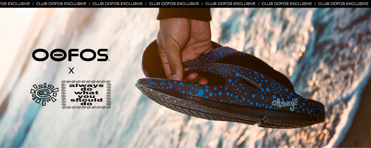 A close-up of a hand holding a black OOFOS flip-flop sandal with bright blue speckled straps and sole details, set against a blurred beach background. The OOFOS logo and a collaboration graphic with 'always do what you should' are visible on the left.