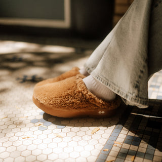 A person wearing tan fuzzy slip-on slippers with white socks and light-wash jeans, standing on a sunlit hexagonal tile floor.