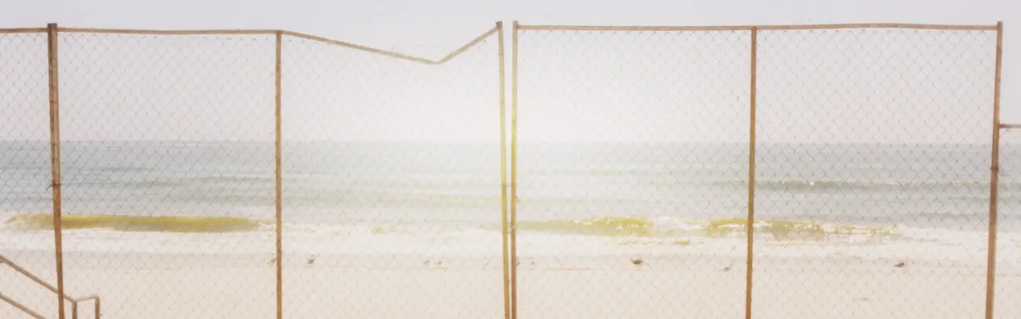 A chain-link fence with a rusty metal frame stands in front of a beach scene with pale sand, calm water, and a bright hazy sky.