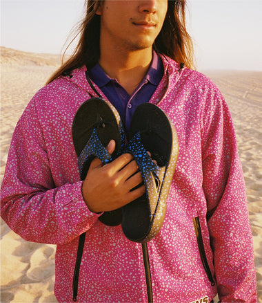 A man in a pink patterned hooded jacket holding a pair of black flip-flops with blue patterned straps, standing in a sandy desert landscape.