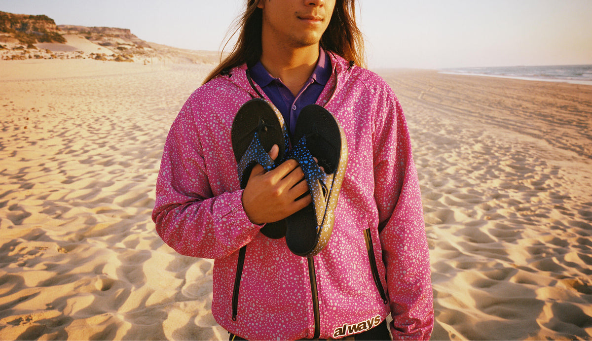 A man standing on a sandy beach wearing a pink patterned zip-up hoodie and holding a pair of black flip-flops with blue straps.