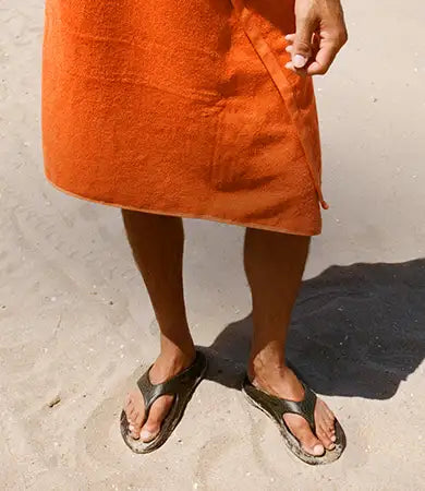An orange beach towel wrapped around the lower half of a masculine person standing on sand, wearing OOFOS × Adsum OOriginal Limited Sandals.
